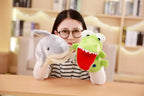 Person holding two plush toys, a gray dolphin and a green dinosaur with a red mouth, in front of a bookshelf.